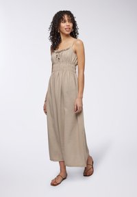 Abito maxi beige con spalline sottili, scollo a volant, vita elastica e spacchi laterali. Modella che indossa sandali flat e accessori minimal.