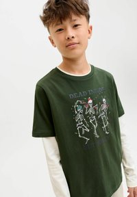 Jeune personne avec les cheveux courts portant un t-shirt vert par-dessus une chemise à manches longues blanche, affichant des squelettes dansants et le texte "Mort à l'intérieur mais joyeux."
