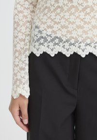 Blonde top med bølgende kant i creme med blomster mønstre, parret med højtaljede sorte bukser. Tekstureret, semi-gennemsigtigt materiale.