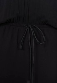Tessuto nero con superficie testurizzata, dotato di una vita elasticizzata e un laccio, creando una forma arricciata in vita.