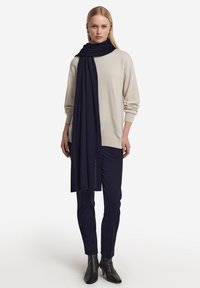 En marineblå uldsjaal hænger over en lys beige sweater, parret med mørke bukser og sorte hæleboots. Tekstur ser blød ud med et subtilt strikmønster.