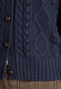 Mørk marineblå kabelstrikket cardigan med store brune knapper over ternede bukser i nuancer af brunt og gult.