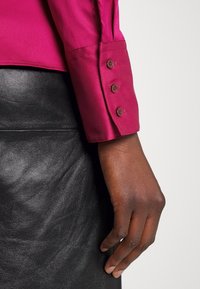 Main à peau foncée reposant près d'une jupe en cuir noir, portant une chemise à manches longues rose foncé avec trois boutons sur le poignet, sur fond blanc.