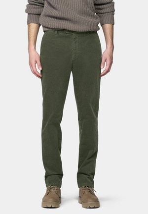 Mann trägt olivgrüne Cordhosen, beige Schnürstiefel und einen braunen Strickpullover mit hochgekrempelten Ärmeln, steht vor einem einfarbigen Hintergrund.
