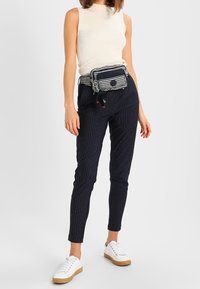 Haut blanc sans manches à côtes, pantalon ajusté rayé bleu marine, et une banane rayée. Le modèle porte des baskets blanches avec des semelles beige.