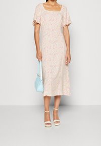 Mujer con un vestido midi floral en tonos pastel y mangas flutter, sosteniendo un bolso azul claro y llevando unos tacones blancos con tiras al tobillo.