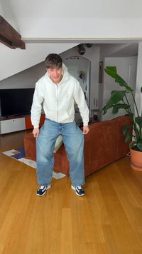 Νεαρός άνδρας που φοράει λευκό φούτερ με κουκούλα και μπλε τζιν σκύβει μπροστά μέσα σε ένα μοντέρνο σαλόνι με καφέ καναπέ και μεγάλο πράσινο φυτό.