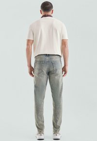 Man die met zijn rug naar de camera staat, gekleed in een wit T-shirt met korte mouwen, lichtblauwe jeans, witte sneakers en een donkere halsdoek, tegen een effen achtergrond.