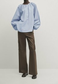 Blouse oversize bleu clair avec manches bouffantes et patte de boutonnage, associée à un pantalon cargo marron ample et des sandales noires.
