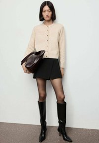 Cárdigan beige de punto con botones dorados, falda corta negra y botas altas de cuero negro. Sostiene un bolso de cuero marrón oscuro.