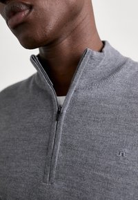 Pull gris avec un col zippé, en tissu maille fine. Dispose d'un petit logo sur la poitrine ainsi que d'un col et d'un ourlet côtelés.