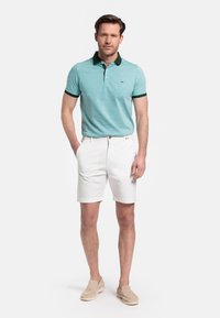 Turquoise poloshirt met donkergroene kraag en manchetaccenten, gecombineerd met witte shorts en beige instappers. Glad stoftextuur.