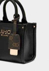 Sac à main en cuir texturé noir avec quincaillerie dorée, doté de doubles poignées et d'une étiquette décorative avec un détail à motif.