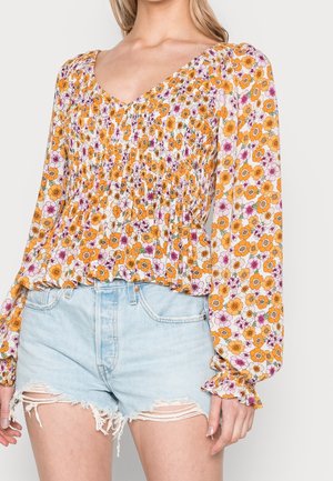 Femme portant un chemisier smocké à manches longues à fleurs orange et violet accompagné d'un short en denim bleu clair déchiré.