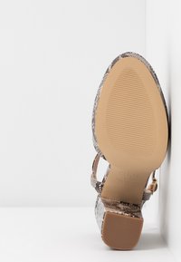 Chaussure à talon haut imprimée lézard positionnée avec la semelle vers l'avant, montrant une semelle extérieure beige texturée et un talon carré.