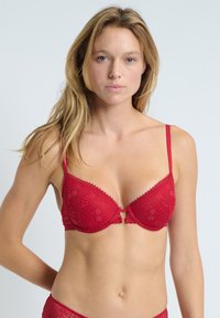 Červená krajková podprsenka s květinovým vzorem, vycpanými košíčky a nastavitelnými ramínky. Uprostřed zdobný otvor ve tvaru klíče. Hladká textura.