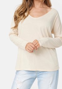 Beige långärmad topp med V-ringning och sänkta axlar, kombinerad med ljusblå slitna jeans. Mjuk tyg med en jämn textur.