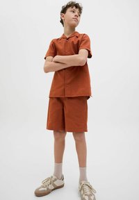 Orange texturerad kortärmad skjorta och matchande shorts, med en campkrage, bärs med beige låga sneakers och ljusa strumpor.
