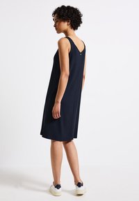 Abito a coste blu navy con schiena a V, senza maniche, lungo fino al ginocchio, e un piccolo dettaglio con bottone sul retro, abbinato a sneaker bianche.