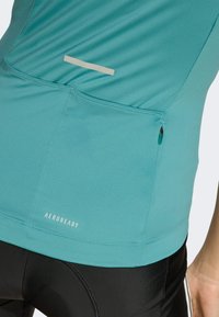 Aqua blue athletic top with textured fabric, a small reflective stripe, and a zippered pocket. Brand logo "AEROREADY" at the bottom.