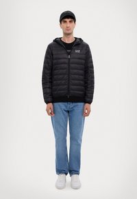 Giacca a vento nera con zip, cappuccio e logo EA7. Indossata sopra una maglietta nera e abbinata a jeans azzurri chiari e sneakers bianche.
