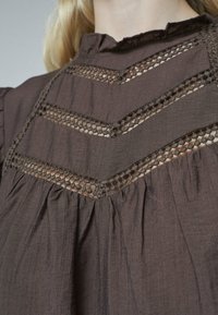 Brun tekstiltop med høj hals, dekorative blonder-lignende udskæringer og lagdelte plisseringer på forsiden for ekstra tekstur.