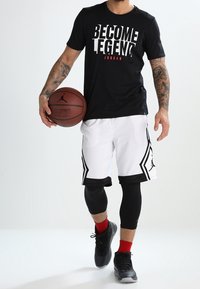 Mann, der ein "BECOME LEGEND"-T-Shirt trägt, weiße Basketballshorts anhat, einen Basketball in einer Hand hält und mit einem Bein leicht nach vorne steht.