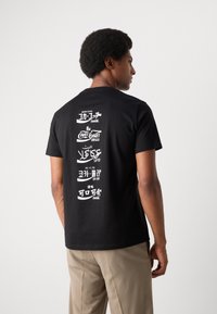 Black cotton t-shirt featuring white text graphics in various languages printed vertically on the back. Short sleeves and crew neckline.