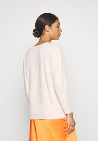 Helles, rosa, flauschiges Strickoberteil mit langen Ärmeln und Rundhalsausschnitt. Der Rücken ist etwas länger als die Vorderseite. Dazu wird ein orangenes Satinkleid kombiniert.