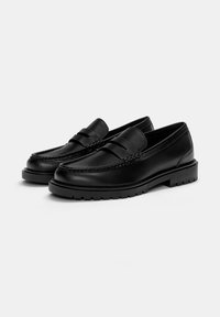I mocassini in pelle nera presentano un design cucito, una suola piatta con piccole scanalature e una fascia decorativa che attraversa la parte superiore.