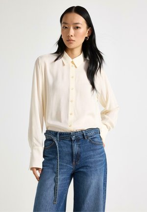 Ung kvinde med langt sort hår iført en cremefarvet skjorte med knapper og blå jeans, stående med den ene hånd i lommen mod en ensfarvet baggrund.