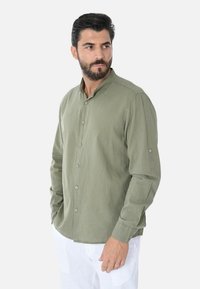 Camicia verde oliva a maniche lunghe con collo alla cinese, bottoni frontali e polsini arrotolati. Realizzata in un tessuto leggero e strutturato.