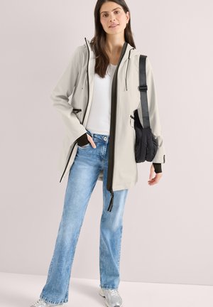 Femme portant une veste à capuche beige, une chemise blanche, un jean bleu clair, des baskets argentées et un sac à bandoulière noir, posant devant un fond uni.