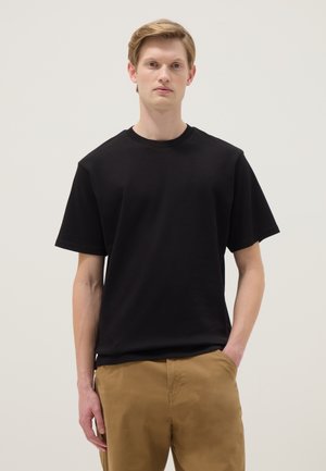 Jonge man die een effen zwarte korte mouwen T-shirt en lichtbruine broek draagt, staat met één hand in de zak tegen een effen achtergrond.