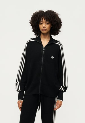 Donna con capelli ricci che indossa una giacca nera Adidas con zip e pantaloni abbinati con dettagli a tre strisce bianche su maniche e gambe.