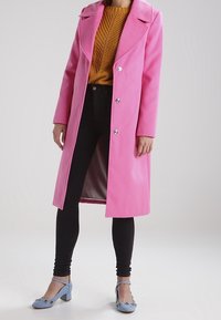 Helles rosa Langmantel mit modernen Revers und doppelten Knöpfen, getragen über einem senfgelben Strickpullover, kombiniert mit schwarzen Leggings und blauen Absatzschuhen.