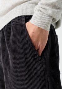 Ruka se odmara u džepu crnih manšestrastih hlača u kombinaciji sa svijetlosivim rebrastim džemperom.