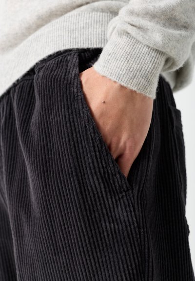 Main reposant dans la poche d'un pantalon en velours côtelé noir porté avec un pull gris clair côtelé.