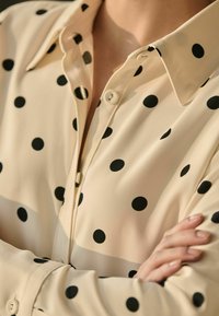 Blouse beige clair avec des pois noirs, manches longues et col à boutons ; fabriquée en tissu lisse avec des détails de couture visibles.