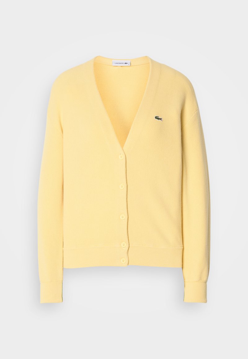 Gelbe Strickjacke mit V-Ausschnitt, Knopfverschluss und gerippten Bündchen. Besitzt ein kleines grünes Logo auf der linken Brustseite. Weiche Textur.