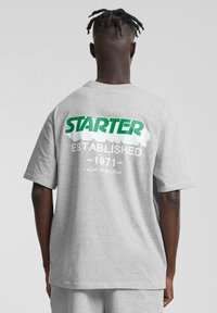 Grå t-skjorte med korte ermer som har en stor grønn "STARTER"-logo, hvit tekst "ETABLERT 1971" og "Se etter stjernen" detaljer på baksiden.