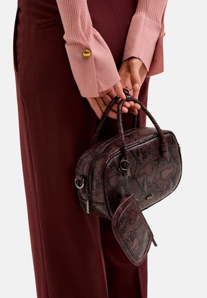 Brązowa i burgundowa torebka w wężowy wzór z górnymi uchwytami, zamek błyskawiczny i dołączona mała kieszonka. Nosi ją w połączeniu z miękkim różowym swetrem i spodniami.