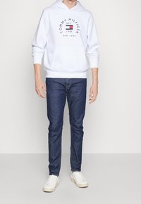 Bílá mikina s kapucí s modro-červeným logem, v kombinaci s tmavě modrými džínami a bílými teniskami. Texturovaná látka, ležérní design.