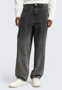 Mężczyzna w luźnych, ciemnoszarych dżinsach z dużymi przednimi kieszeniami, białych trampkach i białej koszuli z długim rękawem.