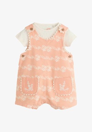 Persikanväriset lyhyet haalarihousut, joissa on aaltoilevat reunat ja puiset napit, valkoisen, ohuella silmukkakuvioinnilla varustetun lyhythihaisen paidan päällä.