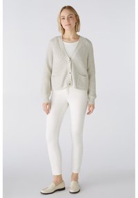 Helle graue Strickjacke aus strukturiertem Material mit V-Ausschnitt, Vorderstaschen und Knöpfen. Kombination mit weißen, eng anliegenden Hosen und Slippern.