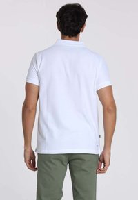 Polo shirt blanc à manches courtes avec un col classique, texture lisse et coupe droite. La vue arrière montre un logo minimaliste à l'ourlet.