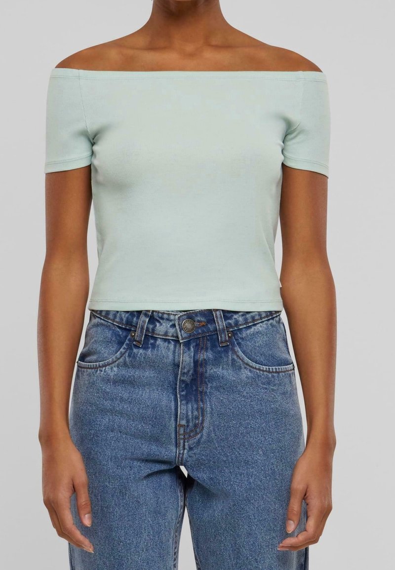 Femme portant un haut court bleu clair à épaules dénudées associé à un jean en denim bleu taille haute, debout devant un fond uni.
