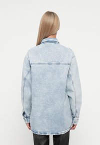 Osoba s dlouhými blond vlasy, oblečená v lehkém modrém oversize denimovém kabátu a černých kalhotách, stojící zády k jasně bílému pozadí.