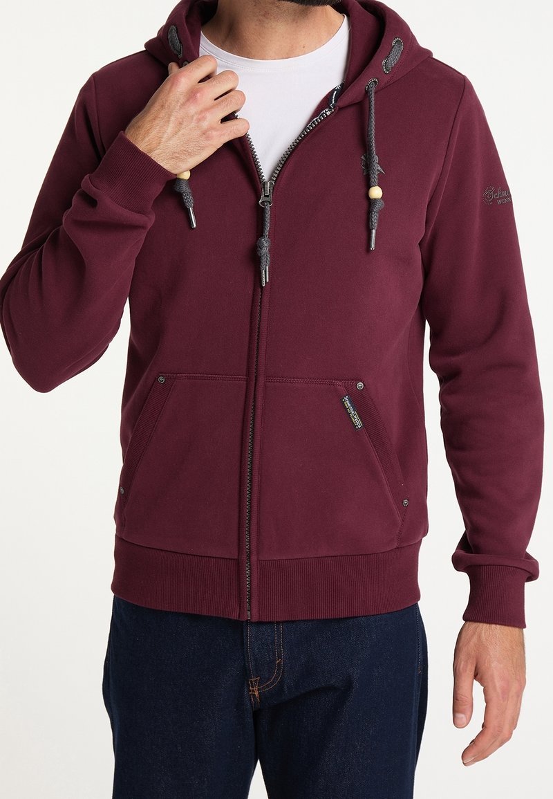 Sudadera con cremallera de color burdeos, con capucha ajustable mediante cordón, dos bolsillos frontales y puños acanalados. Presenta acentos de cuentas de madera en los tiradores de la cremallera.